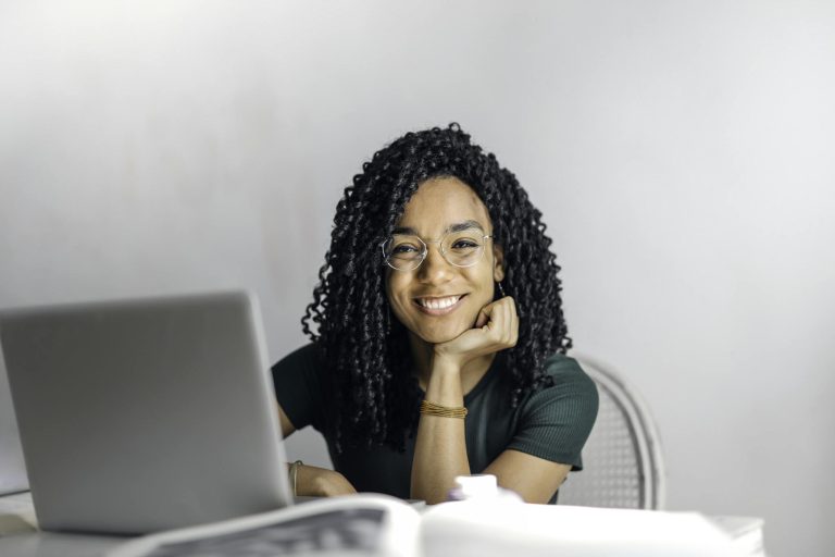 Joyful businesswoman with curly hair smiling at camera while using laptop indoors.